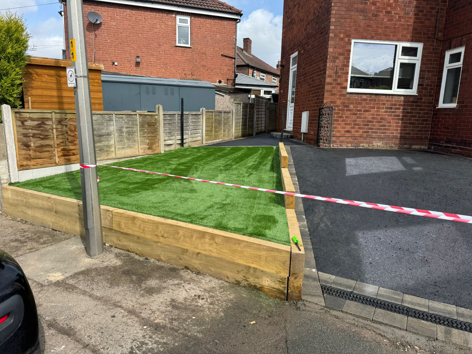 Tarmac driveway beside raised artificial grass retained with timber sleepers